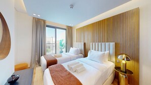 Habitació d'hotel amb dos llits, un finestral gran, una planta en test i una làmpada moderna.