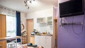 Quarto compacto com cama, escrivaninha, cozinha compacta e televisão na parede.