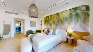 Una habitación de hotel moderna con una cama grande, una zona de estar con una silla, una mesita pequeña y una pared con un mural de hojas de palmera.