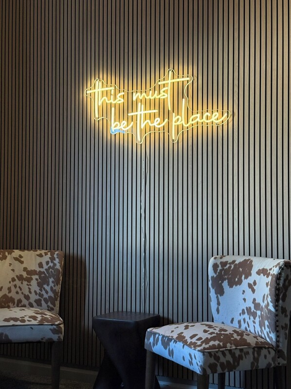 Ein Zimmer mit einer Neonreklame, auf der 'this is the place' steht, einer Holzvertäfelung, zwei Stühlen mit Kuhfellmuster und einem kleinen schwarzen Tisch.