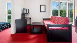 Une pièce avec une moquette rouge, un canapé noir recouvert d’un tissu à motifs rouges, un meuble TV noir et une grande fenêtre donnant sur un paysage verdoyant.