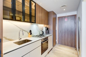 A compact kitchen with a sink, stove, and microwave, wooden cabinets, and a door with a sign. - Sophisticated Studio l Free Pool and Gym (Dubai)