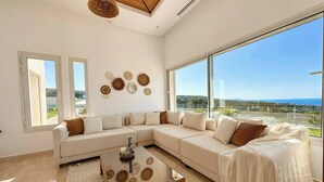 A modern living room with a sectional sofa, a coffee table, and large windows offering a view of the landscape. - Azura House Sea View Pool (Metline)