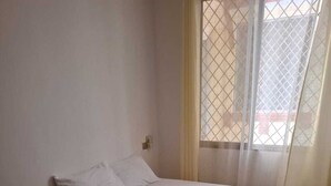 Ein Zimmer mit Einzelbett, einer Matratze, Kissen, einem zusammengerollten Handtuch, einem gemusterten Teppich und einem Paar Flip-Flops auf dem Boden.