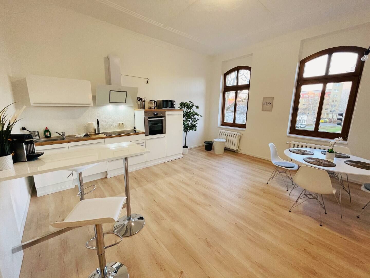 Una cocina moderna con armarios blancos, horno empotrado y microondas. Hay una zona de comedor con una mesa y sillas blancas, y un ventanal que ofrece vistas al exterior.