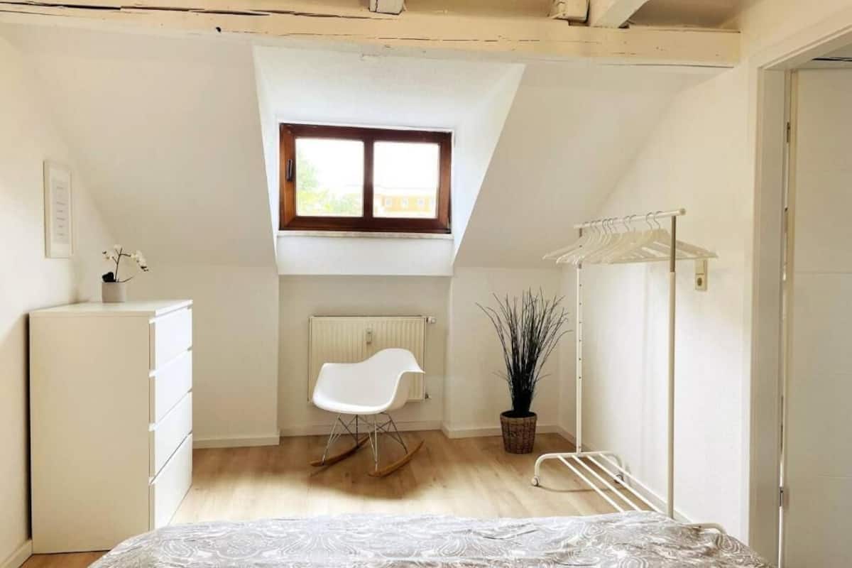 Un dormitorio con techo inclinado, una ventana, un radiador, un aparador blanco, una mecedora, una planta en maceta y un tendedero.