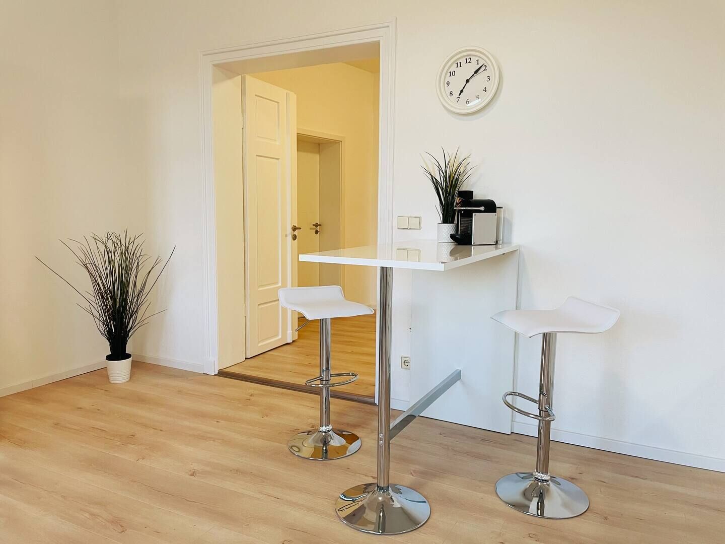Une cuisine moderne avec des meubles blancs, un plan de travail blanc, deux tabourets de bar, une horloge et une plante en pot.