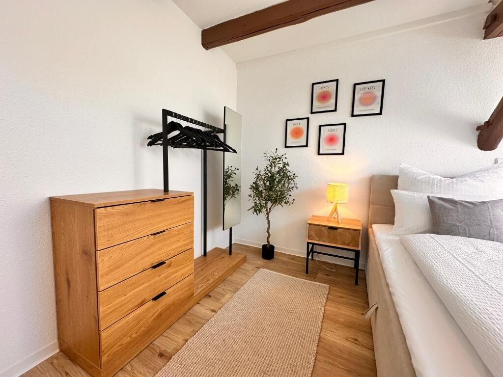 Un dormitorio con un aparador de madera, una mesita de noche con lámpara, una cama con almohadas, un perchero en la pared y cuadros enmarcados.