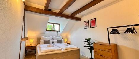 A bedroom with a bed, bedside lamps, a dresser, a plant, and a clothing rack.