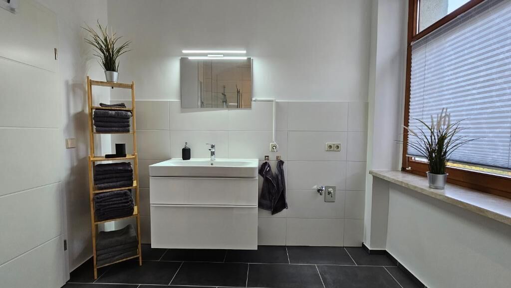 Un baño moderno con tocador blanco, un espejo sobre el lavabo, toallero, estante de madera con toallas y ventana con persianas.