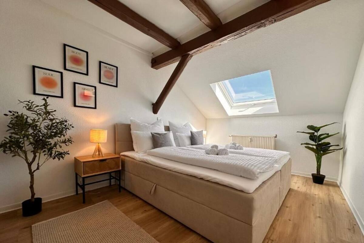 Un dormitorio con techo inclinado, tragaluz, cama con almohadas, mesita de noche con lámpara y una planta en maceta.