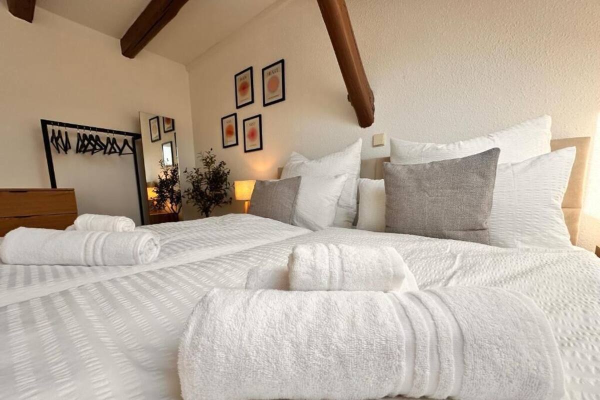 Una cama bien tendida con ropa blanca y toallas enrolladas, un cabecero de madera y un espejo colgado en la pared.
