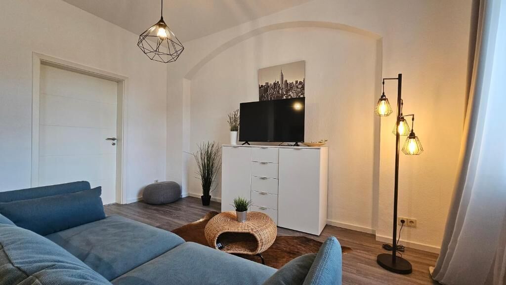 Un saló modern amb un sofà, una televisió, un moble d'estantàries, una làmpada de peu i una obra d'art amb un paisatge urbà.