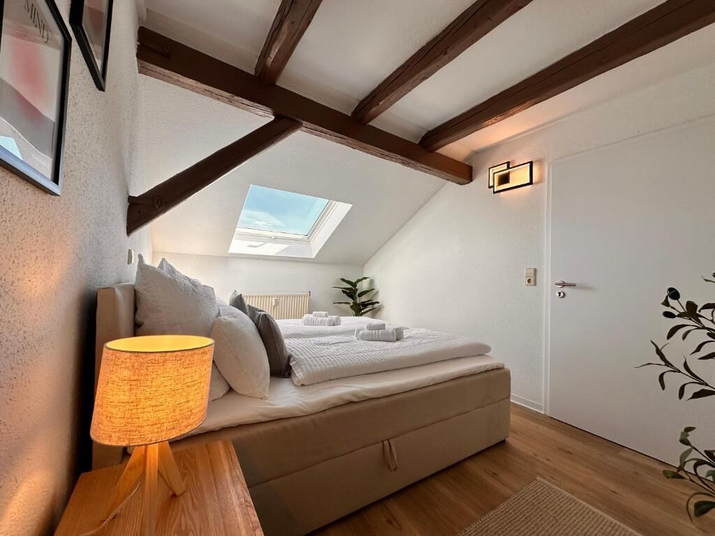 A bedroom with a skylight, a bed with white linens, a bedside lamp, and a wooden floor.
