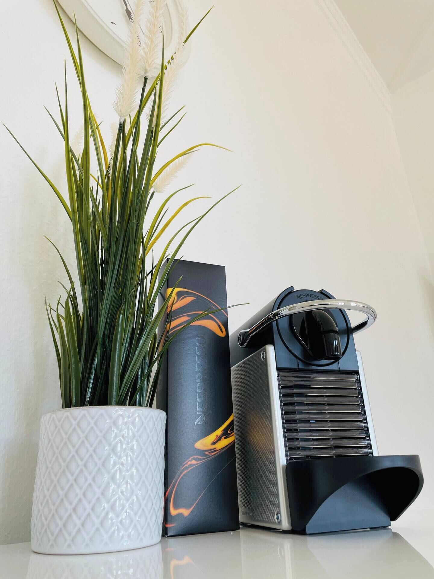 Ein Blumentopf, eine orange-schwarze Box und eine schwarz-silberne Espressomaschine auf einer spiegelnden Oberfläche.