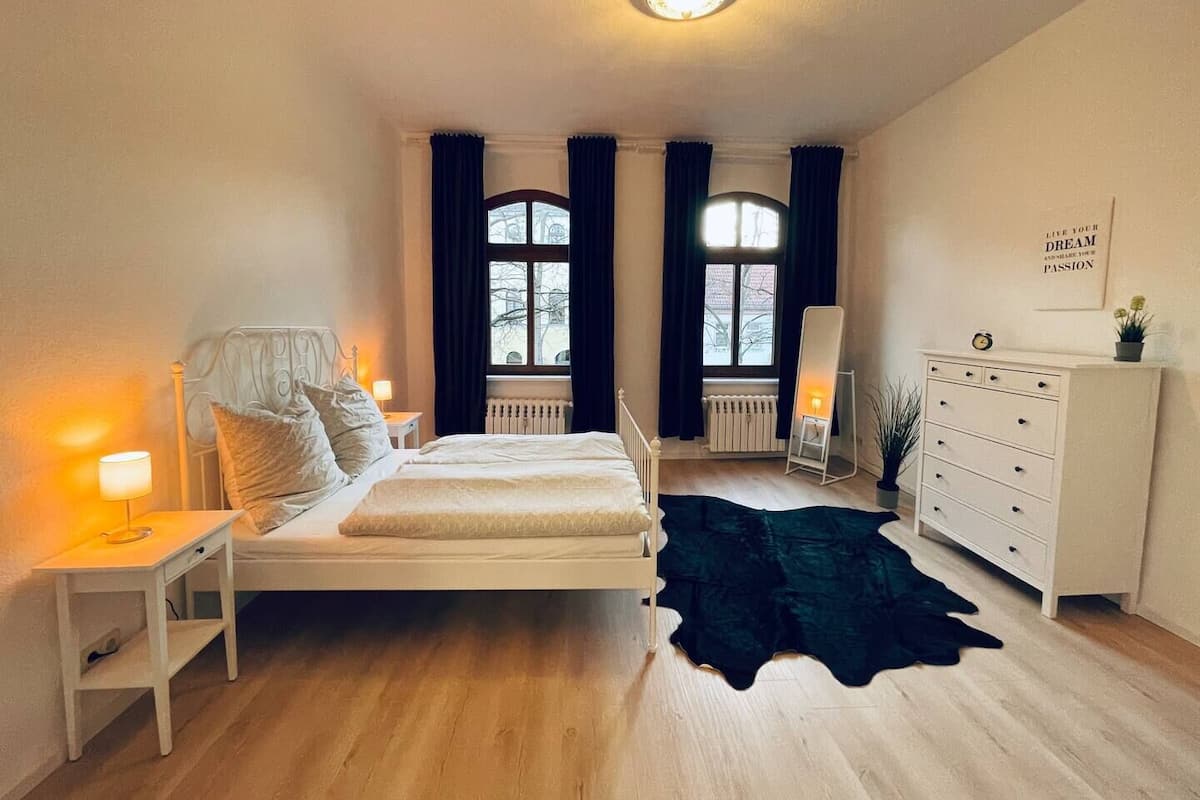 Un dormitorio con cama, mesitas de noche, cómoda, radiador y alfombra negra.