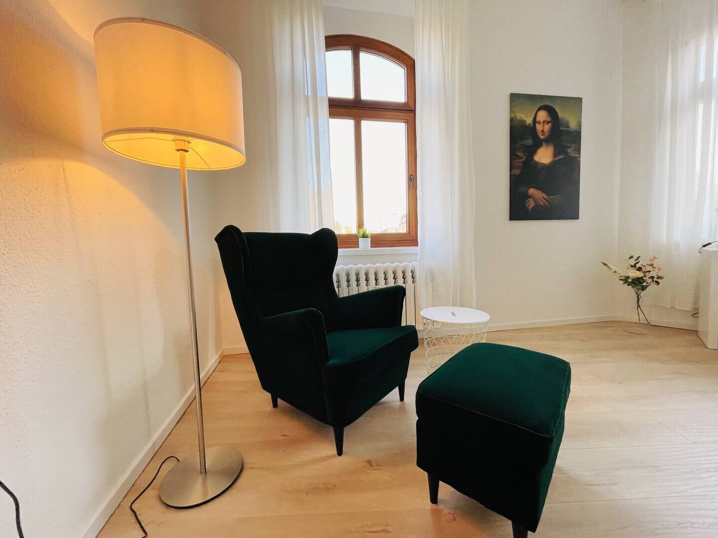 Ein Zimmer mit Stehlampe, Sessel, Hocker, einem Fenster mit Vorhängen, einer Heizung und einem Bild an der Wand.