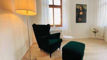 Ein Zimmer mit Stehlampe, Sessel, Hocker, einem Fenster mit Vorhängen, einer Heizung und einem Bild an der Wand.