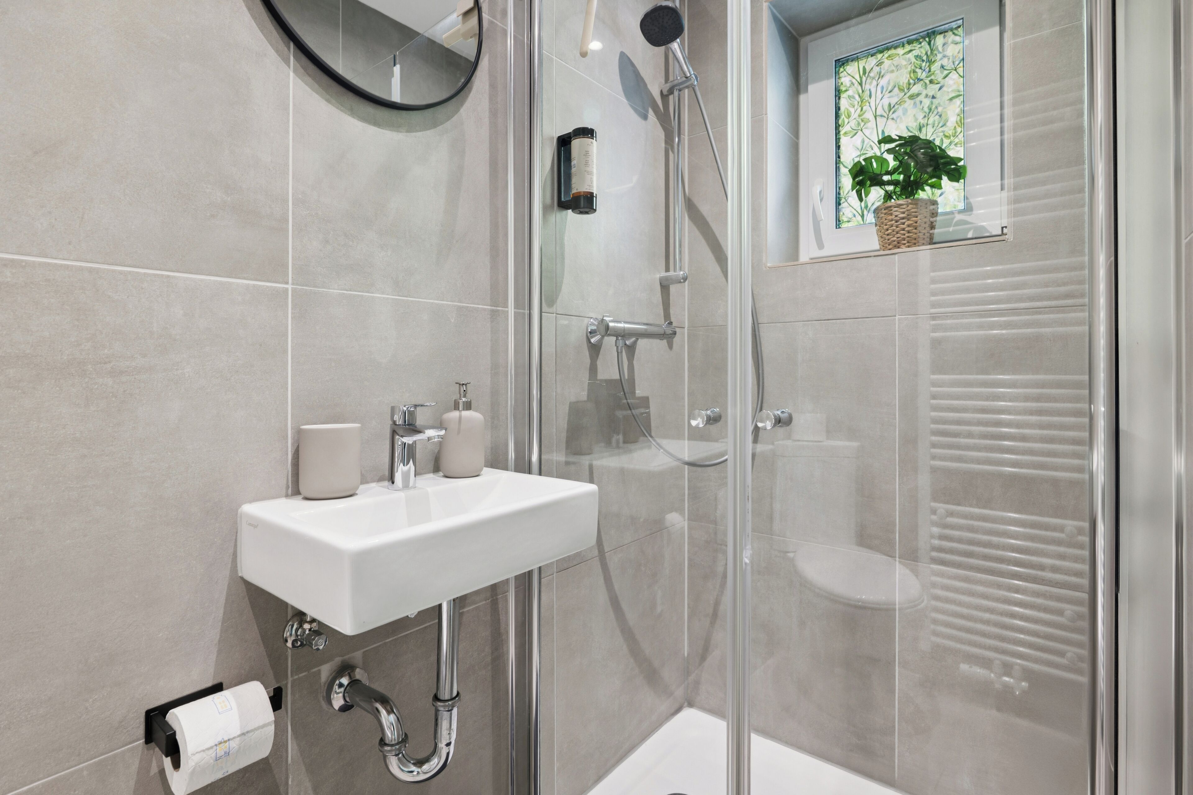 Une salle de bain moderne avec une douche en verre, un lavabo blanc et une petite fenêtre agrémentée d’une plante.