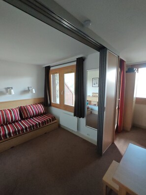 A room with a sofa, a table, a window with curtains, and a door leading to another room. - Le Nid des Silènes – In the Heart of the Cimes 1800 (La Plagne Tarentaise)