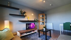 A living room with a TV, shelves with plants, a wooden door, and a small table. - 2BR- Modern, top floor apartment  -  Free parking fast internet.  Near hospitals (Aberdeen)