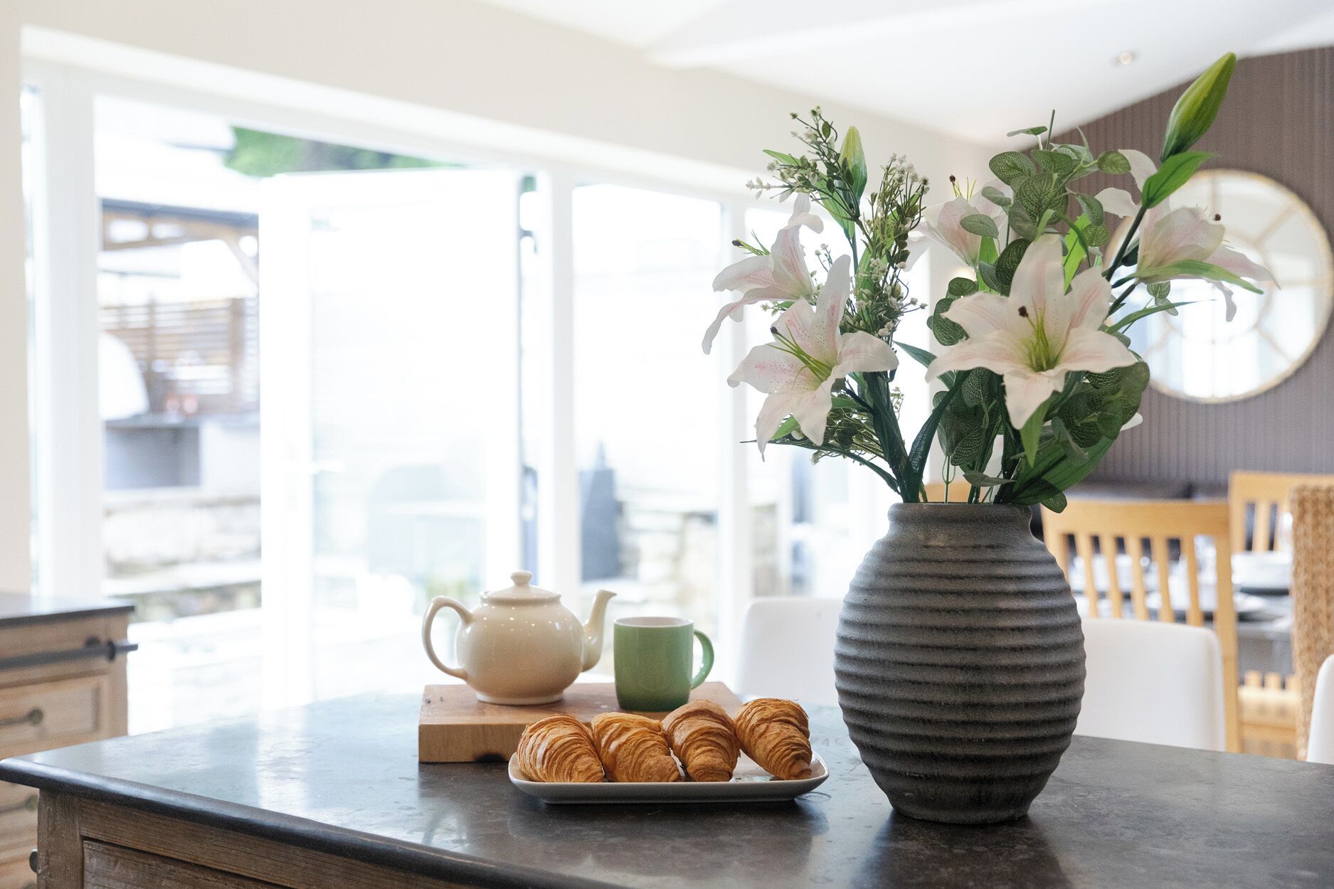 A kitchen counter with croissants, a teapot, a green mug, and a vase with flowers.