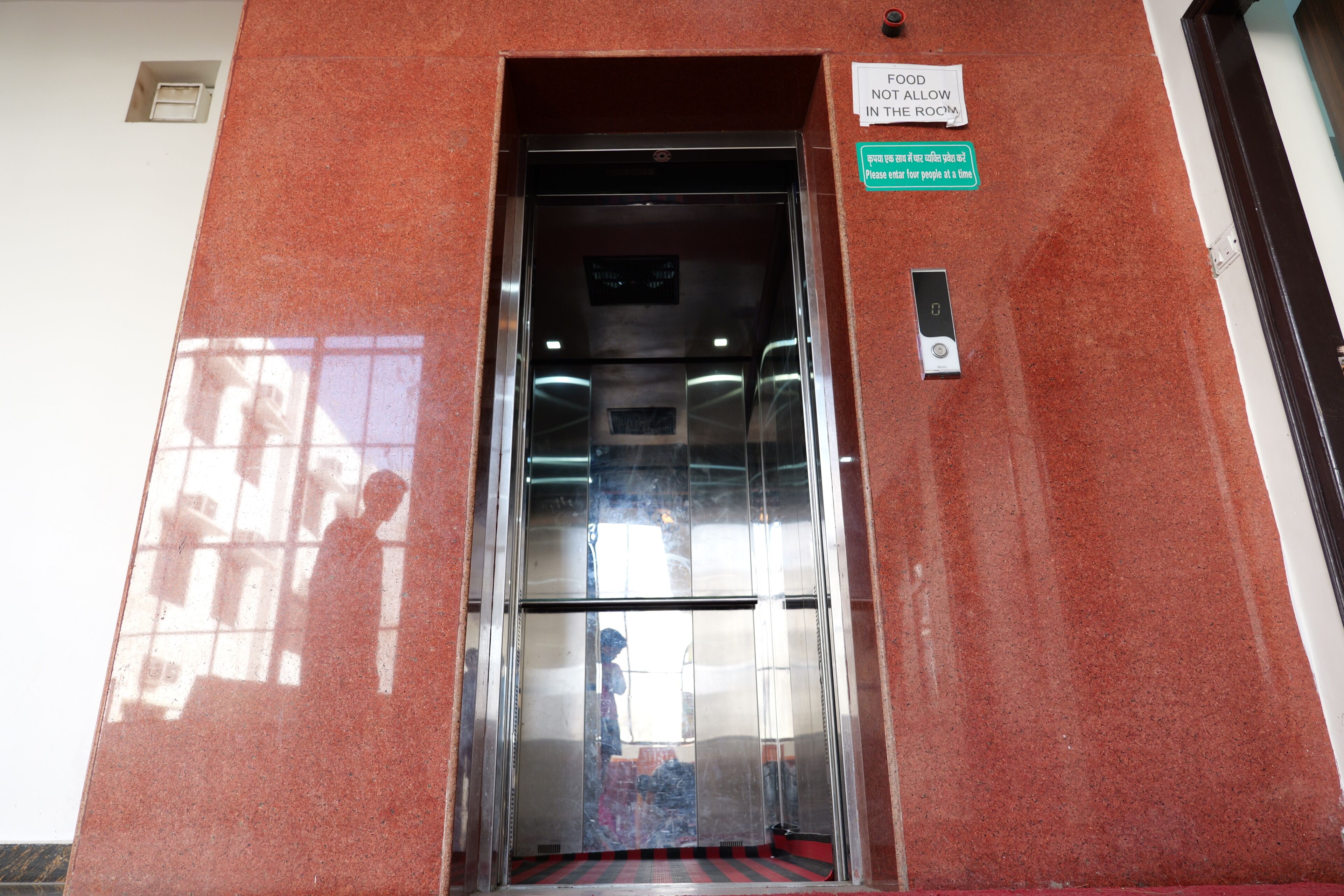 Un ascensor con un letrero que indica 'No se permite comida en la habitación' y un mensaje en otro idioma solicitando que entren menos personas a la vez.