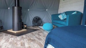A room with a wood-burning stove, a fan, a blue sofa, and a blue bedspread. - Geodome with Hot Tub (Dorset)