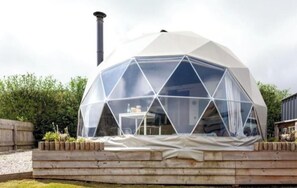 A geodesic dome structure with a chimney, surrounded by a wooden deck and greenery.