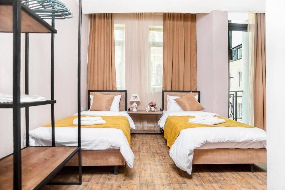 Habitación doble con dos camas, suelo de madera, ventana con cortinas y una pequeña mesa con lámpara.
