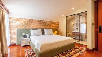 A hotel room with a brick wall, a bed with white linens, a nightstand with a lamp, and a glass shower enclosure.