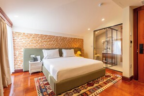 A hotel room with a brick wall, a bed with white linens, a nightstand with a lamp, and a glass shower enclosure.