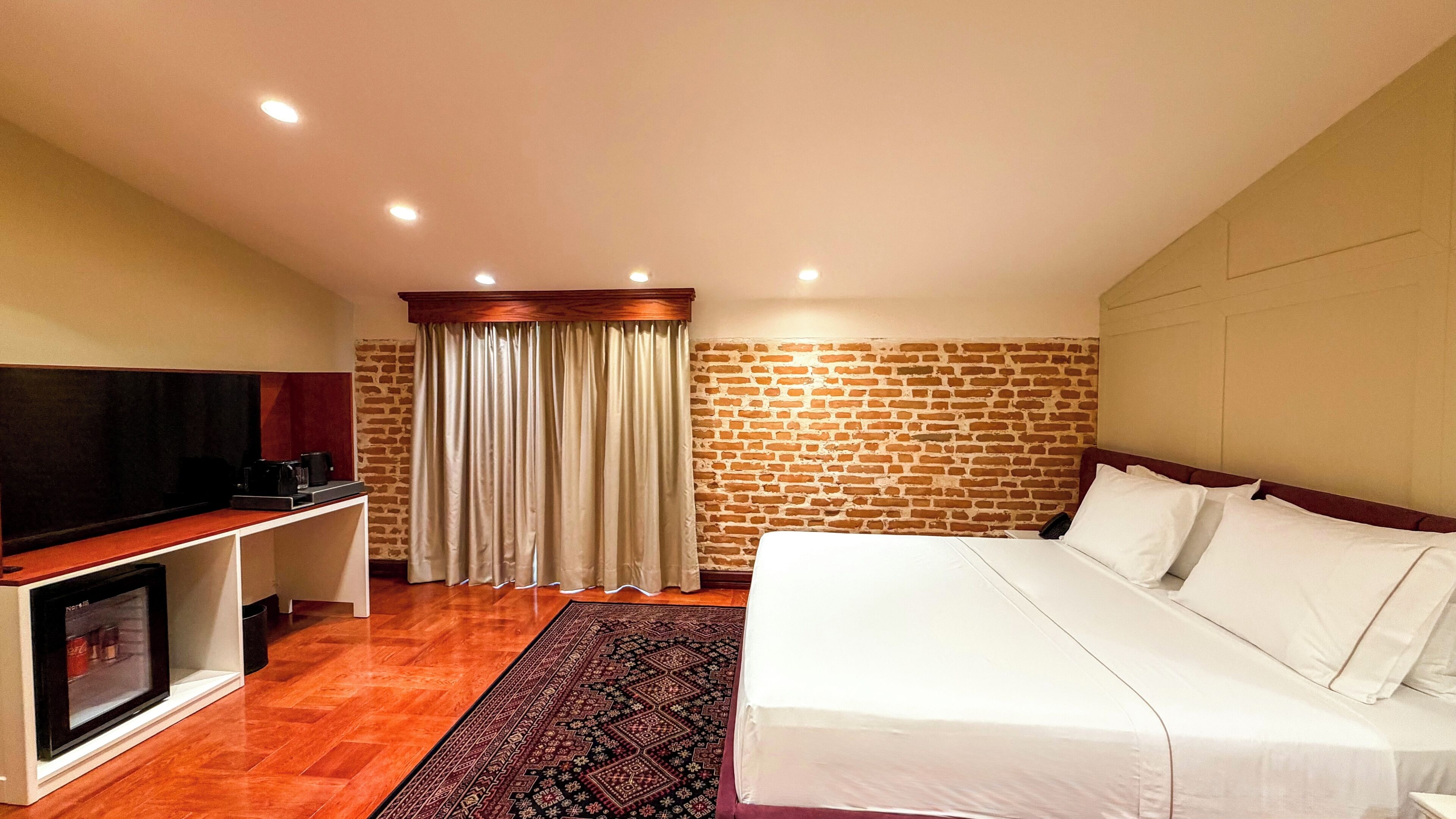 Habitación de hotel con cama, televisión, cafetera, pared de ladrillo y cortina.
