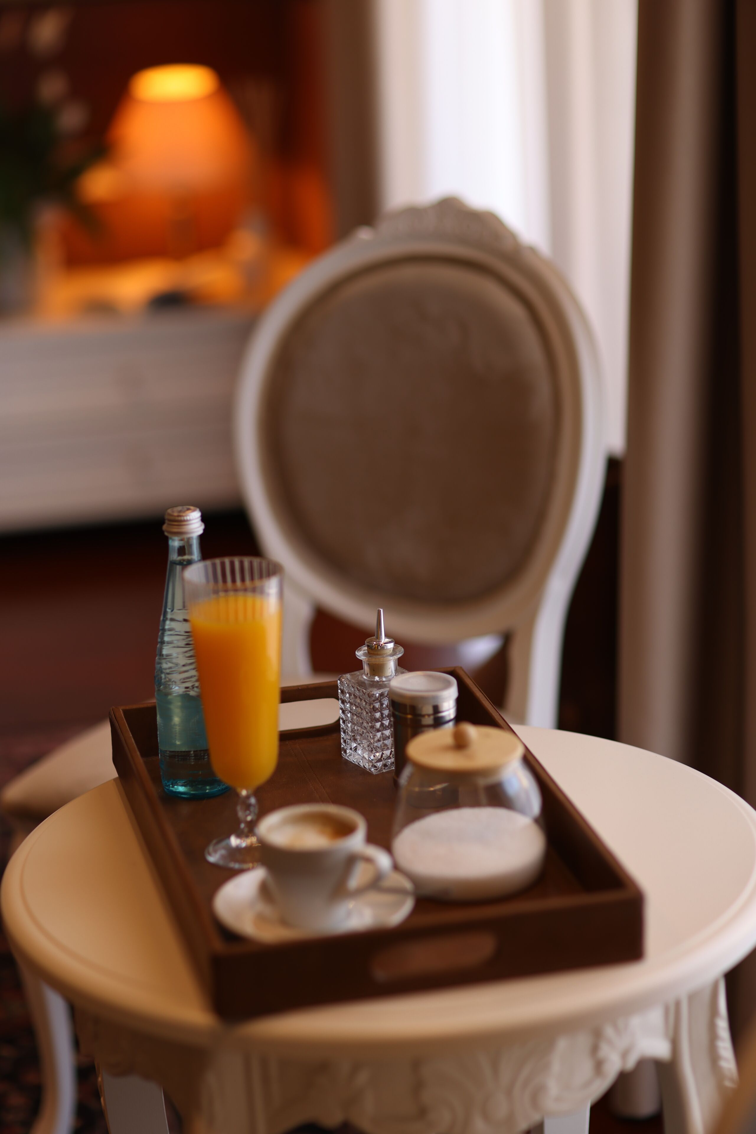 A breakfast tray with a glass of orange juice, a cup of coffee, a bottle, and a small jar on a table in a room with a chair and a lamp in the background.