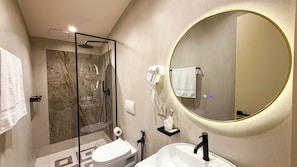 A modern bathroom with a glass-enclosed shower, a round mirror, a white sink, and a toilet. - Vatër Hotel (Shkodër)