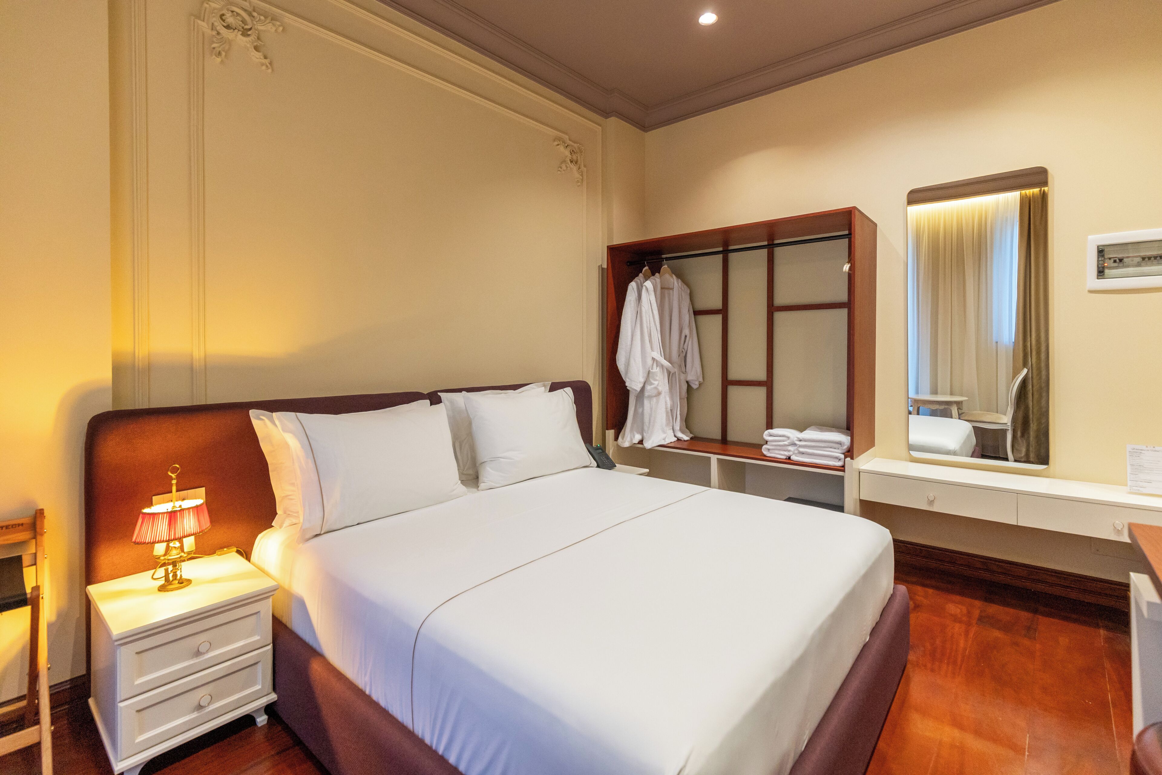 A hotel room with a large bed, a nightstand with a lamp, a mirror, and a robe hanging on a rack.