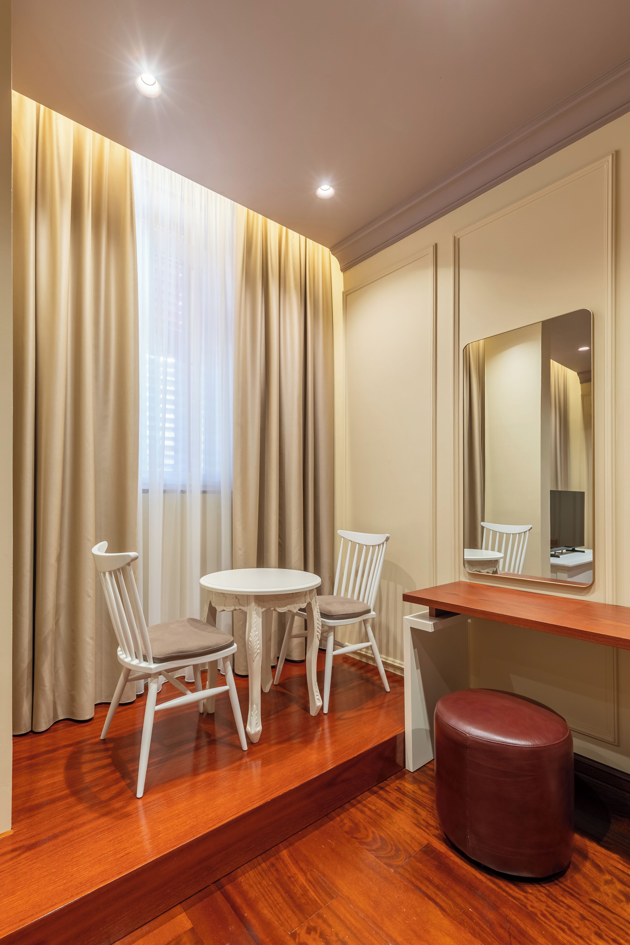 A room with a wooden floor, a round table with chairs, a mirror, and a red ottoman.