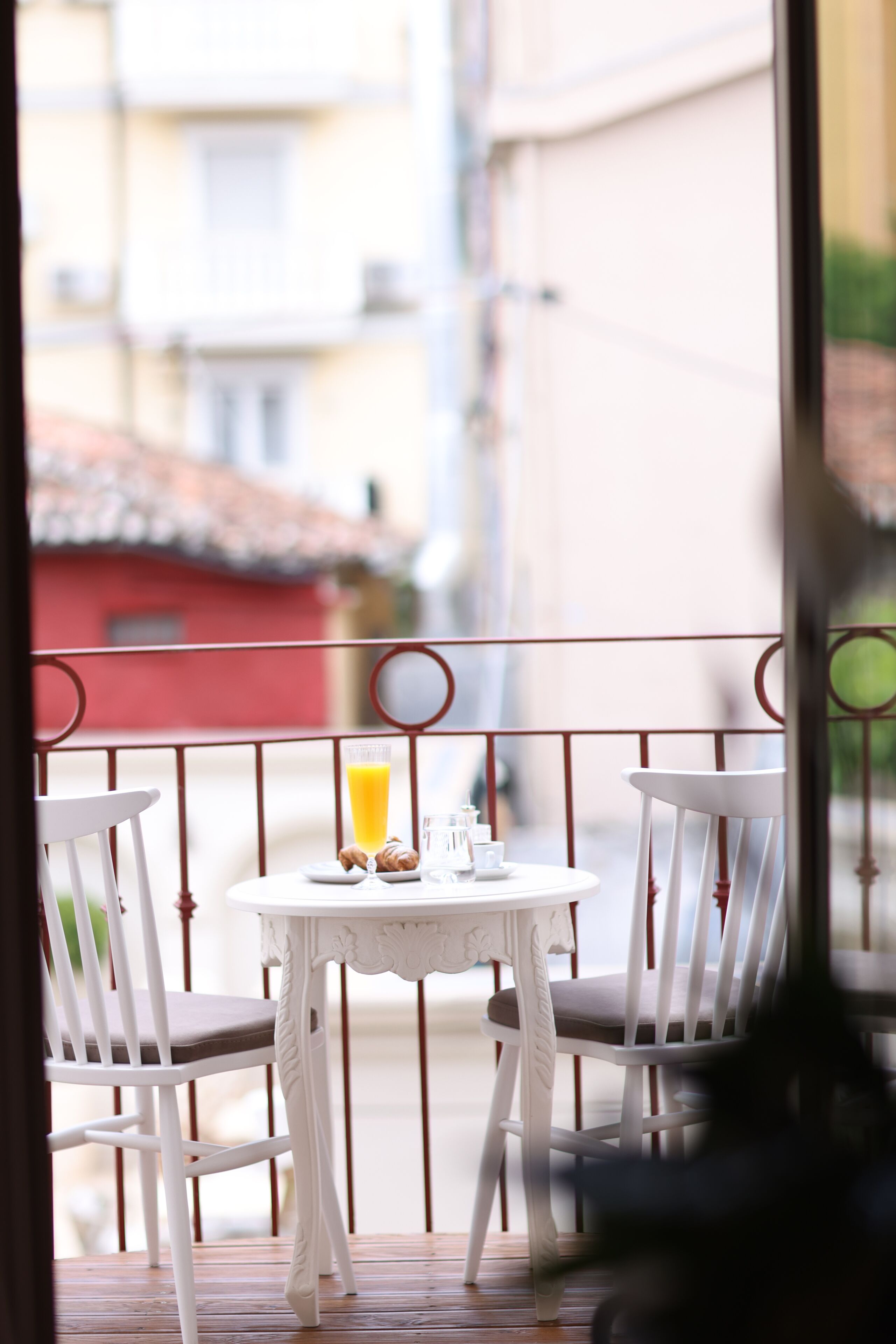 A balcony with a table and chairs, a glass of orange juice, and a red building in the background.