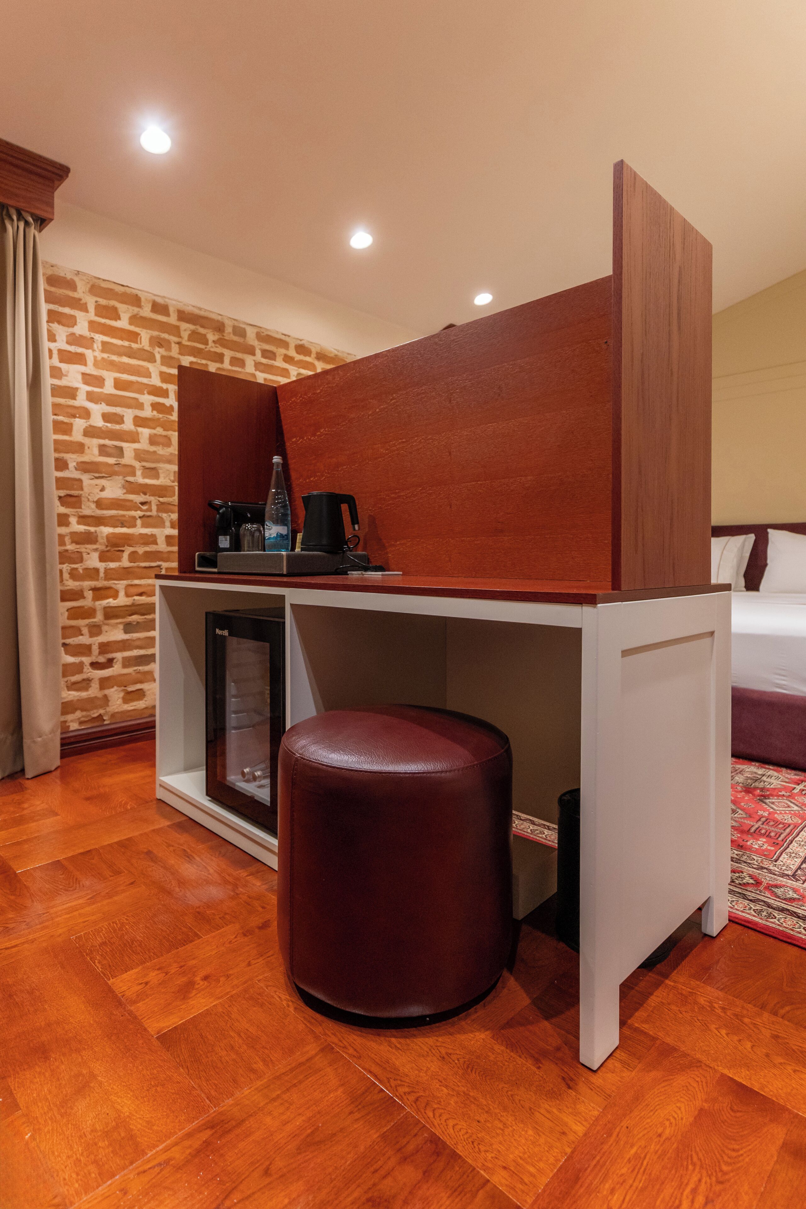 Une chambre d’hôtel avec un bureau en bois, un tabouret rembourré, un mur décoré d’un motif en briques et un lit recouvert d’une couvre-lit à motifs.