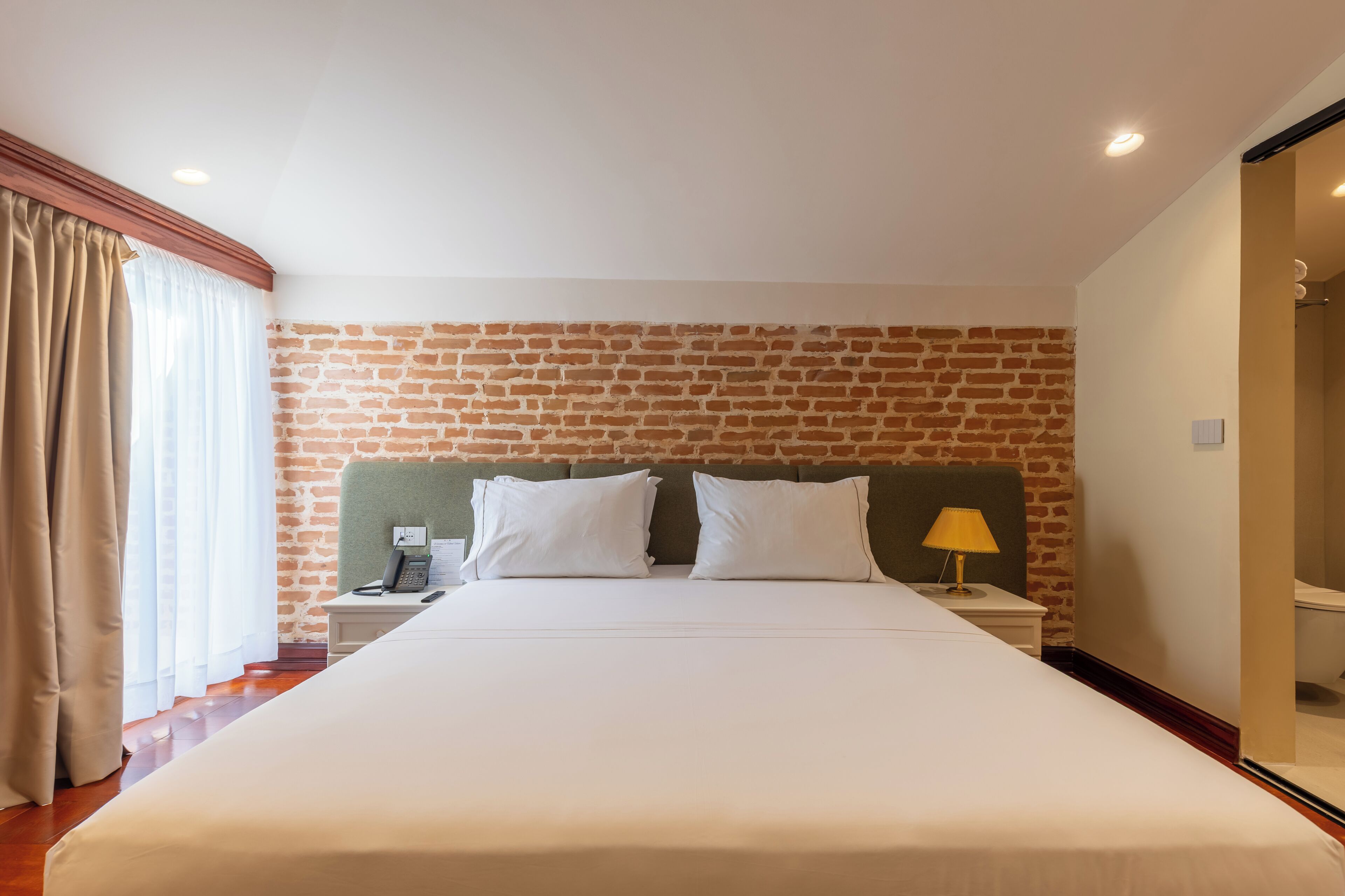 Habitación de hotel con pared de ladrillo, cama con ropa blanca, mesita de noche con lámpara y baño visible a través de una puerta abierta.