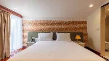 Habitación de hotel con pared de ladrillo, cama con ropa blanca, mesita de noche con lámpara y baño visible a través de una puerta abierta.