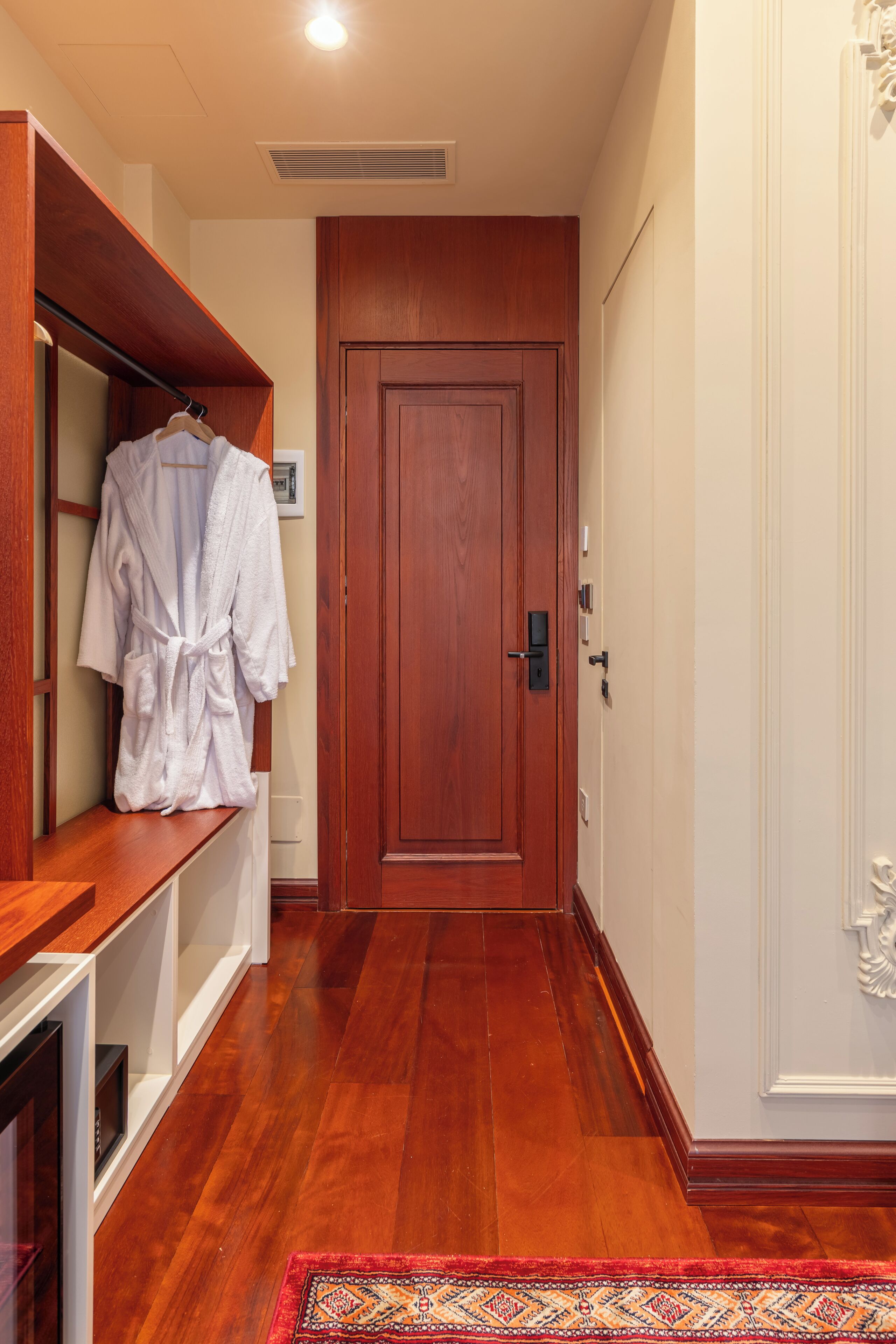 A hallway with a wooden floor, a red patterned rug, a wooden bench with a robe, and a closed door.