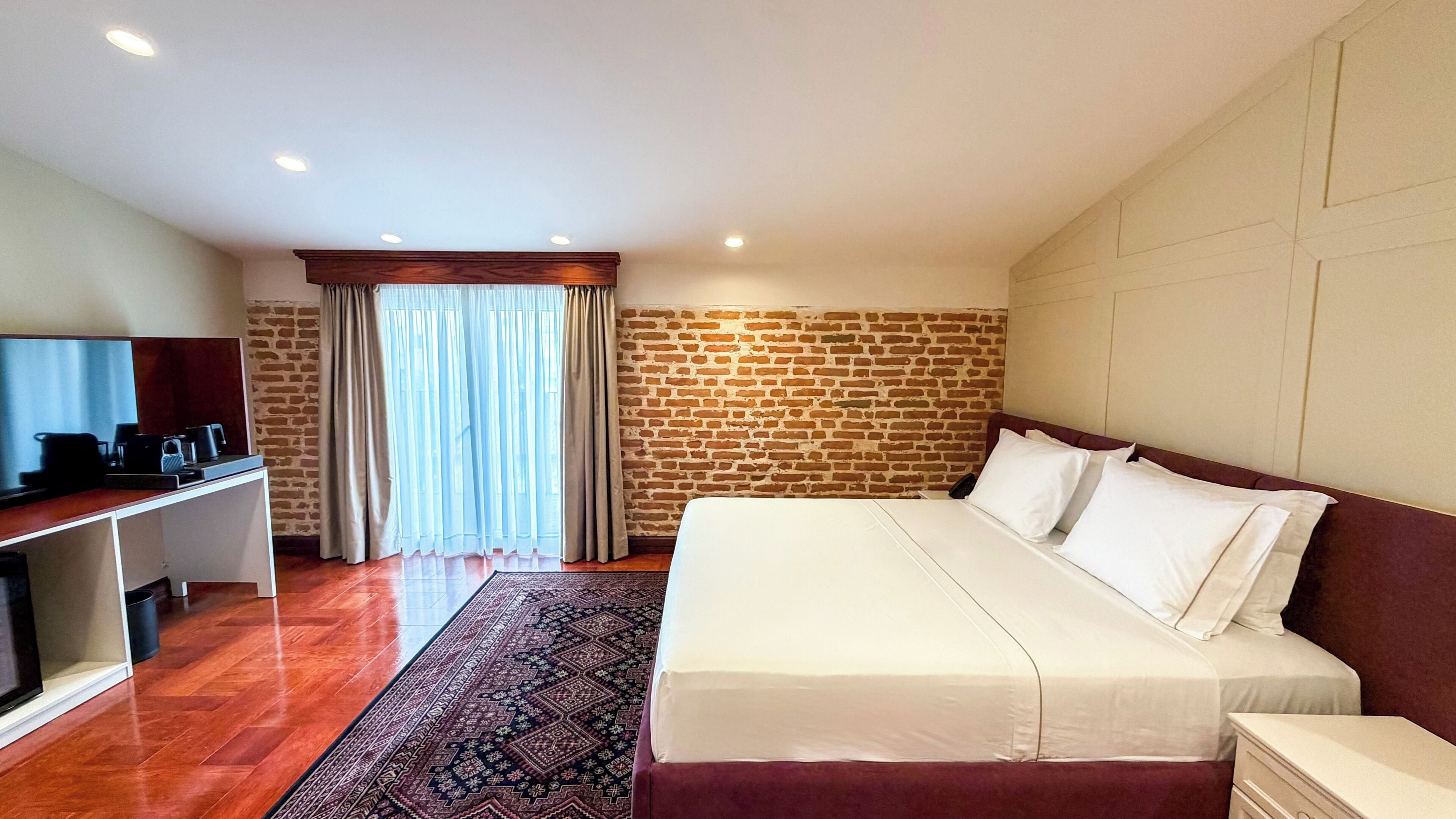 A hotel room with a brick wall, a bed with white linens, a desk with a television, and a patterned rug.
