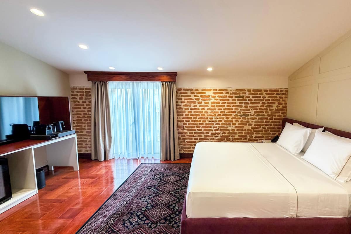 A hotel room with a brick wall, a bed with white linens, a desk with a television, and a patterned rug.