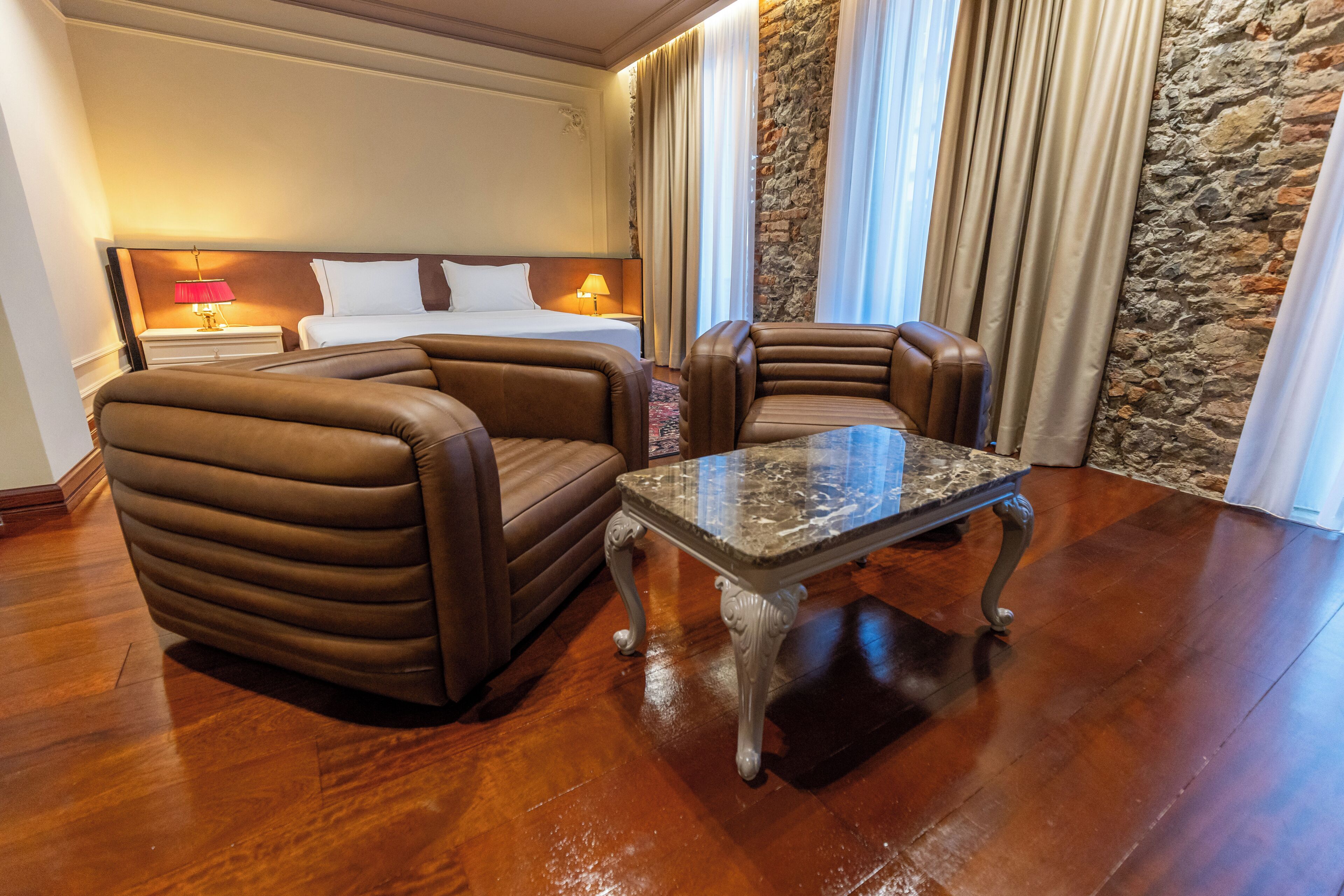 Una habitación de hotel con una cama, dos sillones de cuero, una mesa de centro de mármol, una mesita de noche con lámpara y una pared de acento de piedra.