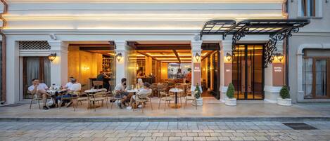 An outdoor cafe with tables and chairs, a bar, and a hotel entrance.