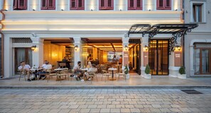 An outdoor cafe with tables and chairs, a bar, and a hotel entrance.