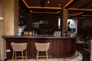 A bar area with a wooden counter, two wicker barstools, and a kitchen in the background. - Vatër Hotel (Shkodër)