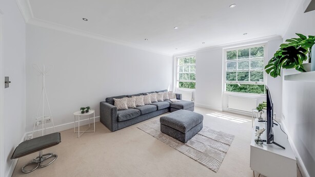 A modern living room with a grey sectional sofa, a grey ottoman, a white coffee table, and a large window with a view of greenery. - The Marylebone Hacienda (London)