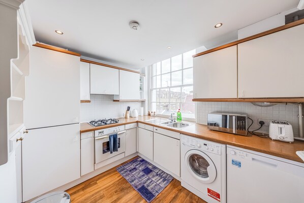 A modern kitchen with white cabinets, wooden countertops, a built-in oven, a microwave, a dishwasher, and a large window. - The Marylebone Hacienda (London)